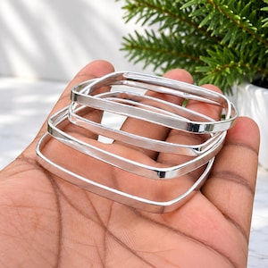 May include: A set of three polished silver-toned square bangles with a smooth, reflective finish. The bracelets feature a distinct four-sided geometric shape with rounded corners, creating a clean and uniform appearance across each individual piece.