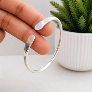 Peut inclure: Un bracelet manchette en métal argenté poli avec une surface lisse et arrondie et une conception ouverte. Le bracelet présente une bande simple et continue qui se courbe en forme circulaire, affichant une finition métallique réfléchissante.
