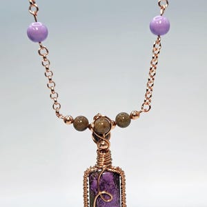 May include: A handcrafted necklace featuring a rectangular purple pendant with a copper wire frame and a decorative swirl. The necklace includes a copper chain, lavender beads, and brown beads. The pendant measures approximately 1 inch in length.