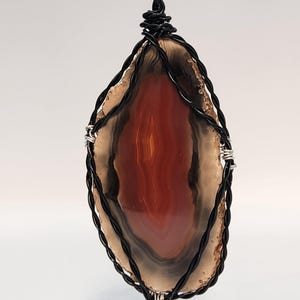 May include: A pendant necklace featuring a polished agate stone with shades of red, brown, and cream. The stone is encased in a black wire wrap with silver accents, suspended from a black chain. The agate has a natural, layered appearance.