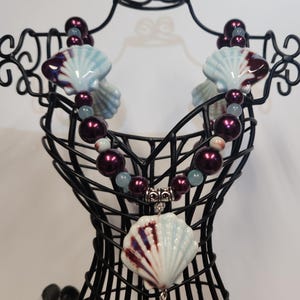 May include: A necklace with a blue and purple seashell pendant and burgundy beads. The necklace is displayed on a black jewelry stand.