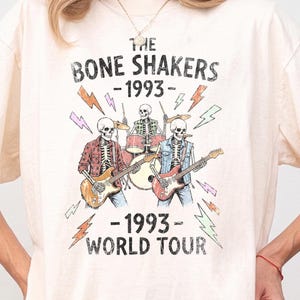 May include: An ivory short-sleeve t-shirt featuring a graphic of three skeletons playing instruments with the text THE BONE SHAKERS 1993 WORLD TOUR. The design includes colorful lightning bolts and a distressed vintage-style print on the front.