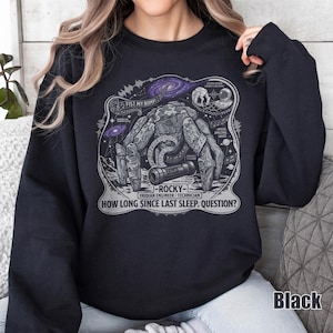 May include: A black crewneck sweatshirt featuring a graphic print of a rocky, multi-limbed creature. The design includes space elements, musical notes, and text reading FIST MY BUMP, ROCKY, ERIDIAN ENGINEER TECHNICIAN, and HOW LONG SINCE LAST SLEEP. QUESTION?