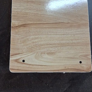 May include: A rectangular board with a light brown wood grain finish and a smooth, reflective surface. The board features four small, evenly spaced circular holes located near each of the four corners, providing a clean and functional design.