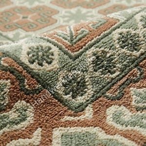 Puede incluir: Una alfombra de pelo insertado con un patrón geométrico en tonos verde salvia, terracota y crema. El pelo grueso y texturizado muestra motivos florales y angulares repetitivos. El texto Handwerker-Teppiche es visible en la superficie.