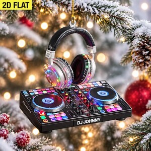 Puede incluir: Un adorno colgante plano en 2D que presenta un controlador de DJ negro y plateado con el texto DJ JOHNNY impreso en el frente, junto con auriculares negros y plateados con detalles de luces de colores, suspendido por un cordón dorado.