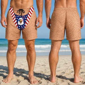 May include: Tan swim shorts with a textured surface. The front features a triangular panel with a blue background, white stars, and red and white stripes. A graphic of a bald eagle wearing sunglasses is centred, surrounded by red and blue fireworks.