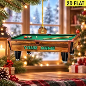 May include: A 2D flat hanging ornament shaped like a pool table with a green felt surface, wooden side panels, black legs, and two cues. The side of the table features the text "YOUR NAME" in green letters, and a gold cord is attached for hanging.