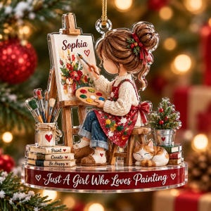 May include: A clear acrylic Christmas ornament featuring a girl painting on a canvas labeled Sophia with a heart. She wears a cream sweater and red apron, sitting on a stool with a cat. The base reads Just A Girl Who Loves Painting.