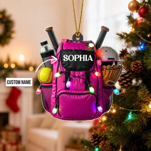 May include: A hanging ornament shaped like a bright pink backpack featuring the name SOPHIA in white text. The backpack is decorated with colorful string lights and holds a yellow softball, a water bottle, a baseball helmet, and a glove.