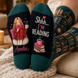 May include: Dark green and black socks featuring a graphic of a blonde person reading a book with the name Amelia printed below. The other sock displays a stack of books, a pink mug, and the text Shhh... I'M READING. The upper portion of the socks features a repeating pattern of books on shelves.