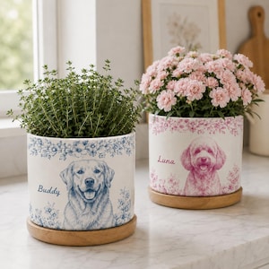 Puede incluir: Dos macetas cilíndricas de cerámica con platos de madera. La maceta izquierda tiene una ilustración azul de un perro con el nombre Buddy. La maceta derecha tiene una ilustración rosa de un perro con el nombre Luna. Ambas con motivos florales.