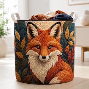 May include: A cylindrical fabric storage basket featuring a quilted exterior with a detailed orange and white fox portrait surrounded by dark blue and golden yellow leaves. The basket includes two white side handles and contains soft items.