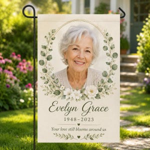 Può includere: Una bandiera da giardino rettangolare con il ritratto di una persona anziana dai capelli bianchi all'interno di una cornice ovale dorata. La cornice è circondata da foglie di eucalipto verdi e fiori bianchi. Il testo recita Evelyn Grace 1948-2023 e Your love still blooms around us.