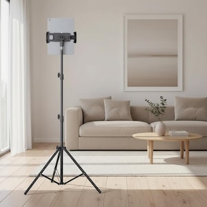 May include: A black metal floor stand with a tripod base and an adjustable telescopic pole, featuring a secure mounting bracket at the top that holds a silver tablet in a vertical orientation for hands-free viewing and stable positioning.