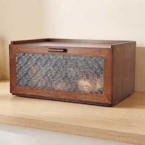 May include: A rectangular wooden bread box with a dark brown finish and a horizontal handle. The front panel features a clear glass window with a decorative embossed clover-like pattern, allowing a partial view of the interior contents.