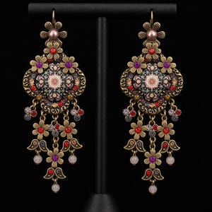 May include: A pair of ornate dangle earrings in an antique gold-tone metal finish. Each earring features a central floral motif with intricate multi-coloured enamel patterns, accented by small red, purple, and clear crystals and dangling flower charms.