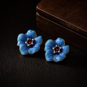 May include: A pair of stud earrings shaped like five-petaled flowers with bright blue enamel petals, gold-toned edges, and a centre cluster featuring small white pearls surrounding a single small red gemstone in the middle of each flower.