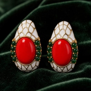 May include: A pair of teardrop-shaped earrings featuring a central oval red cabochon stone surrounded by small green beads. The upper and lower sections of the earrings are finished in white enamel with a gold-toned grid pattern overlay.