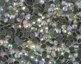 May include: A close-up image of a large quantity of clear iridescent rhinestones. The rhinestones are arranged in a random pattern and are all the same size. The rhinestones are set on a black backing.