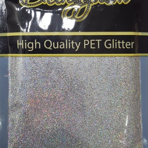 Op de afbeelding: Een doorzichtige plastic zak gevuld met zilveren, iriserende glitter. De zak heeft een zwart label met de tekst "Blinggasm" in gouden letters en de tekst "High Quality PET Glitter" in witte letters. De onderkant van de zak heeft het websiteadres "www.blinggasm.com" in witte letters gedrukt.