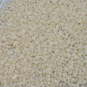 May include: A close-up image of a large quantity of white pearl-like beads. The beads are oval shaped and have a slight iridescent sheen.