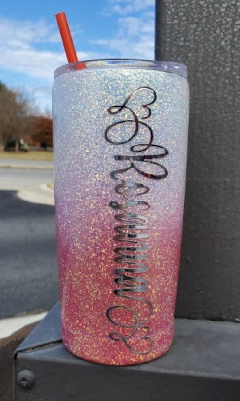 Op de afbeelding: Een roze en witte ombre glitterbeker met een zwarte cursieve inscriptie die "Be Amazing" leest.