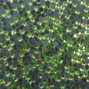 May include: A close-up image of a large quantity of small, round, green rhinestones with a flat back. The rhinestones are scattered on a white surface.