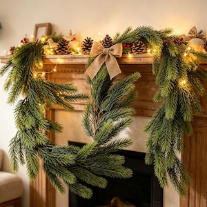 May include: A green artificial pine garland decorated with brown pinecones, red berries, and warm white string lights. A tan burlap bow is centered on the garland, which is draped across a wooden mantelpiece with a dark fireplace opening below.