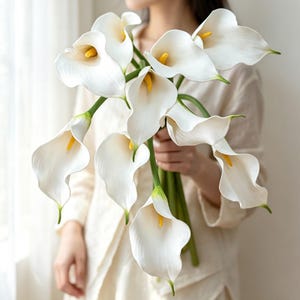 Puede incluir: Un ramo de calas blancas con pétalos lisos en forma de trompeta y espádices de color amarillo brillante. Cada flor está unida a un tallo verde largo y delgado, creando un arreglo natural con formas curvas, elegantes y tonos blancos suaves.