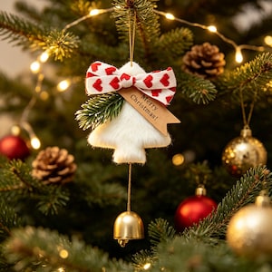 Könnte beinhalten: Ein weißer, flauschiger Weihnachtsbaumschmuck mit einer rot-weißen Schleife mit Herzmuster, einem kleinen grünen Tannenzweig und einem beigen Holzanhänger mit der Gravur Merry Christmas!. Eine goldene Metallglocke hängt an der Unterseite.