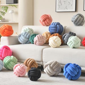 Puede incluir: Una colección de cojines de nudo esféricos en una amplia variedad de colores sólidos, incluyendo naranja, rosa, azul, verde, gris, beige y blanco. Cada cojín está construido con un tubo de tela continuo tejido en una forma de nudo compleja y redondeada.