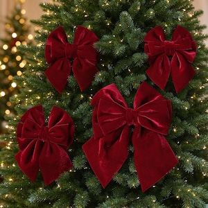 May include: Four deep red velvet bows with multiple layered loops and long, pointed tails. The bows feature a soft, plush texture and are arranged in a cluster, showcasing a rich, dark crimson colour that contrasts with the green pine needles.