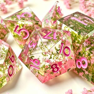 Puede incluir: Un conjunto de dados poliédricos hechos de resina transparente, cada uno con pequeñas flores rosas y follaje verde incrustados. Los números de los dados están grabados y rellenos con un pigmento rosa brillante, creando una estética floral.