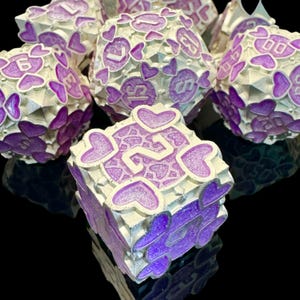 May include: A set of polyhedral dice featuring a silver metallic finish with recessed purple glitter enamel. Each face is decorated with heart-shaped cutouts surrounding the white numbers. The dice set includes various shapes such as a d20, d12, and d6, all displaying a consistent heart-themed design with clear, legible numbering on each face.