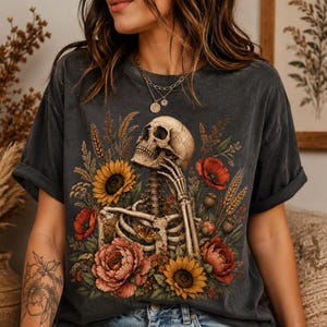 May include: A charcoal gray short-sleeve t-shirt featuring a graphic print of a human skeleton surrounded by vibrant sunflowers, red poppies, and wheat stalks. The skeleton is depicted with one hand resting near its chin amidst the floral arrangement.
