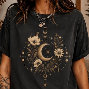 Może przedstawiać: Ciemnoszary t-shirt z krótkim rękawem z centralnym nadrukiem graficznym przedstawiającym złoty półksiężyc w otoczeniu szczegółowych ilustracji kwiatów, liści i małych gwiazd ułożonych w kolisty wzór z delikatnymi geometrycznymi liniami.