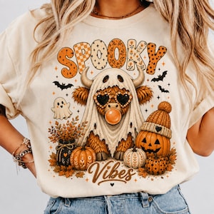 Op de afbeelding: Een crèmekleurig t-shirt met een afbeelding van een hooglandrund in een spooklaken, met hartvormige zonnebrillen en een kauwgombal. Het ontwerp bevat de tekst "SPOOKY Vibes", pompoenen, een spookje, vleermuizen en herfstbladeren.