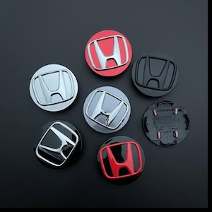 May include: A collection of seven circular Honda wheel center caps featuring the iconic H logo. The set includes various finishes such as chrome, matte black, red with chrome, and silver, with one cap showing the black plastic underside.