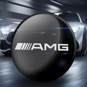 May include: A circular black emblem featuring the silver AMG logo with a stylised graphic element on the left side. The surface is smooth and reflective, displaying the text AMG in a bold, sans-serif font centred on the round component.