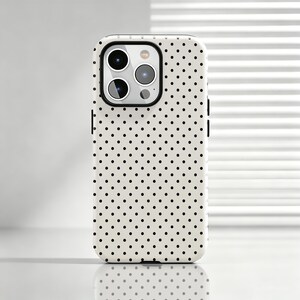 Op de afbeelding: Een smartphonehoesje met een crèmekleurige achtergrond en een herhalend patroon van kleine, gelijkmatig verdeelde zwarte stippen. Het hoesje heeft een rechthoekige uitsparing in de linkerbovenhoek voor een drievoudig camerasysteem.