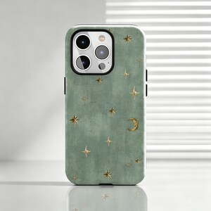 May include: A sage green smartphone case featuring a textured finish with a scattered pattern of gold-coloured four-pointed stars and a single crescent moon. The case has a black camera module cutout with three circular lenses and a flash.