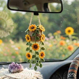 May include: A crocheted sunflower hanging ornament featuring a small brown basket with yellow flowers and green leaves on a jute string, suspended from a car rearview mirror above a cream-colored macrame coaster with a purple crystal cluster.