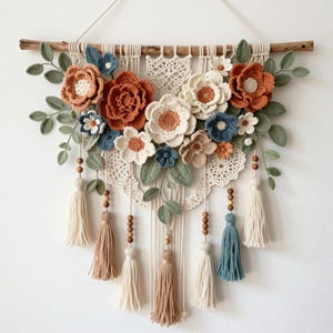 May include: A wall hanging featuring crocheted flowers in shades of rust, cream, and dusty blue, accented with green crocheted leaves. The arrangement is suspended from a wooden branch and includes cream and blue tassels with wooden beads.