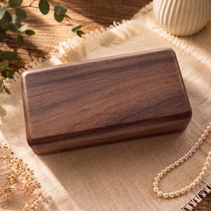 May include: A rectangular wooden box with a smooth, dark brown finish and visible wood grain. The box features rounded edges and a flat, solid lid. The construction is simple and clean, showcasing the natural texture and deep color of the wood.