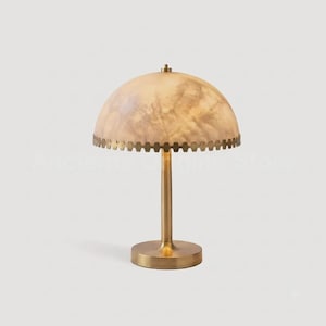 May include: A table lamp featuring a dome-shaped shade with a marbled, cream-colored surface and a decorative scalloped brass edge. The lamp has a slender, cylindrical brass stem and a circular brass base, providing a stable foundation.