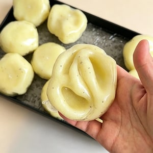 May include: A collection of pale yellow, semi-translucent, rounded wax melts with a smooth, glossy finish and small dark specks embedded throughout. One individual wax melt is held up to show its irregular, folded, and soft-looking surface texture.
