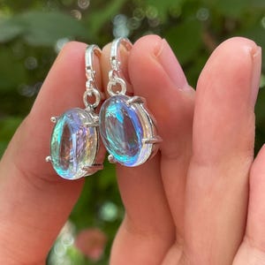 May include: A pair of silver drop earrings featuring oval-shaped iridescent stones. The stones display a translucent quality with shifting blue, purple, and pink reflections. Each stone is secured in a four-prong setting attached to a silver hook.