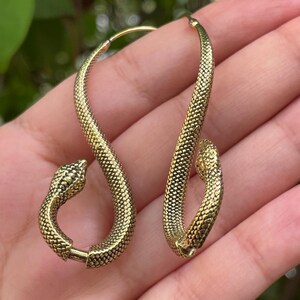 May include: A pair of gold-toned hoop earrings shaped like snakes. Each earring features a textured, scale-like surface throughout the body, with a detailed snake head at one end and a smooth, curved tail section that connects to form a circular hoop.