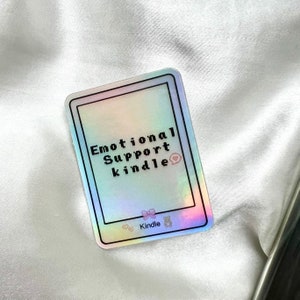 May include: A holographic sticker with the text "Emotional Support Kindle" in black text. The sticker has a pink bow and a pink bear icon. The text "Kindle" is also printed on the sticker.